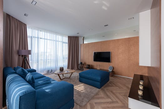 Interior of spacious living room with couch and pouf placed on rug near table and window with curtains against wall with TV
